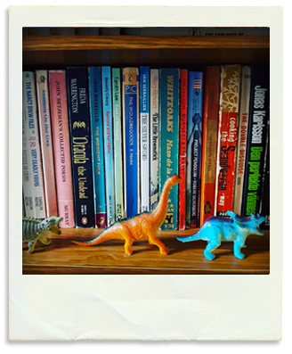 A bookshelf containing vintage paperbacks with three model dinosaurs in front of them.